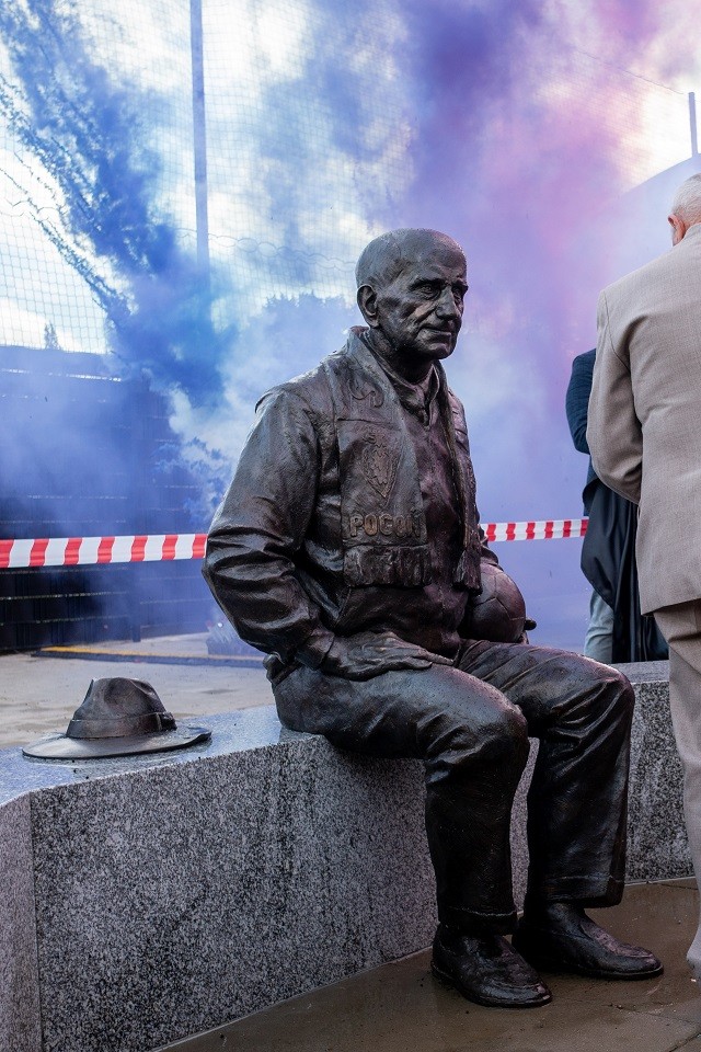 na zdjęciu znajduje się pomnik Floriana Krygiera siedzącego na murku a w tle leci fioletowy dym