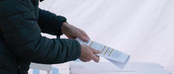 Ilustracja przedstawia mężczyznę w zielonej kurtce wrzucającego kartę do głosowania do urny oznaczonej logiem SBO
